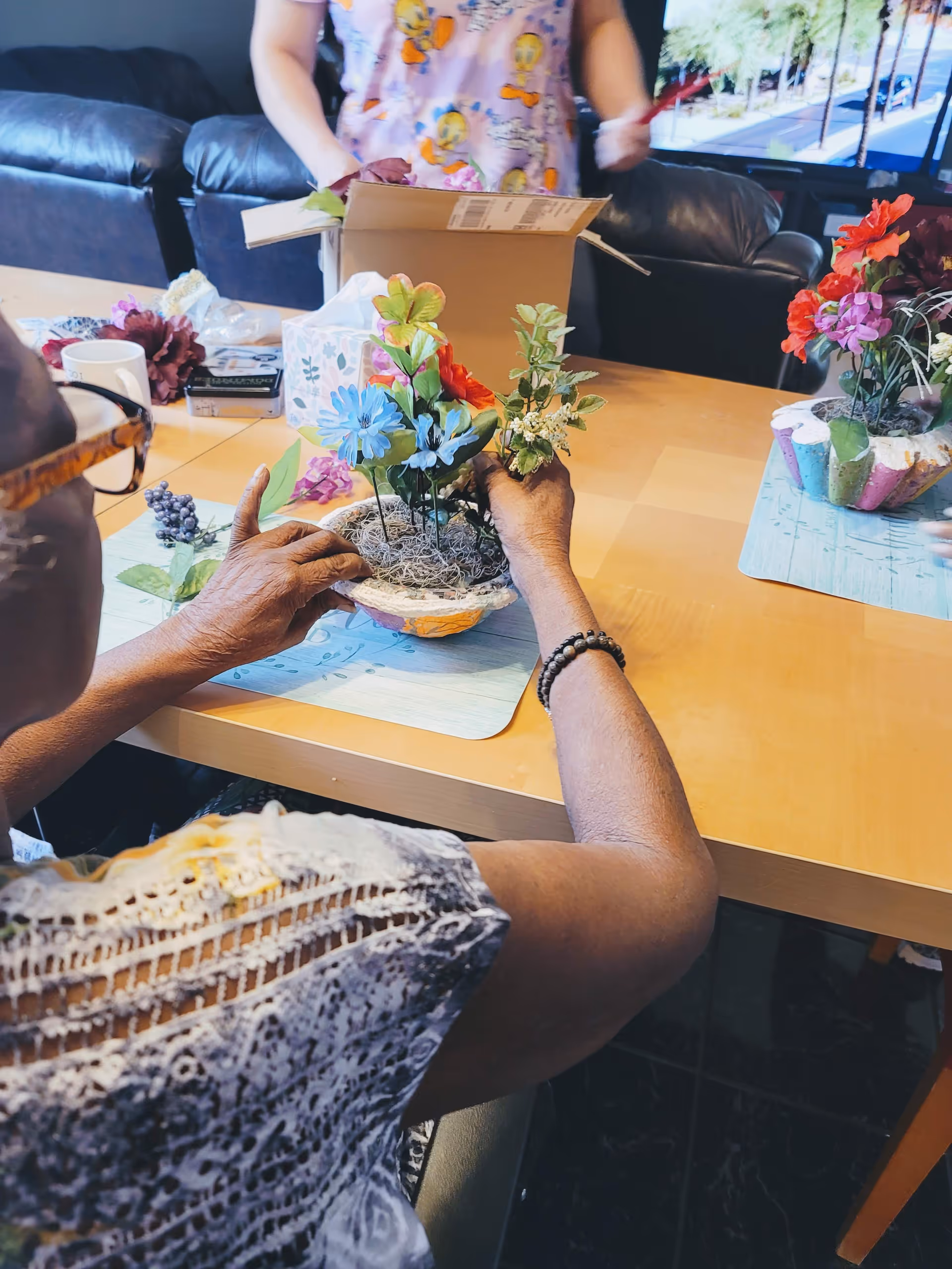 A seated resident arranges a small artificial-flower centerpiece on a table during a group activity with another person and a couch visible in the background.