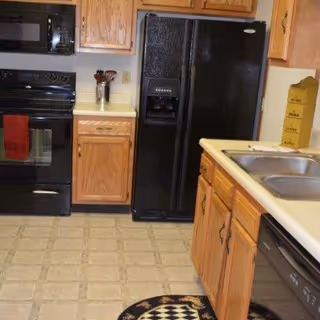 Kitchen area with wooden cabinets, a black refrigerator with water and ice dispenser, a black oven with a red towel hanging on the handle, a microwave above the oven, a double sink, and a dishwasher. The floor has a light-colored tile pattern and a small round rug with a black and white geometric design.