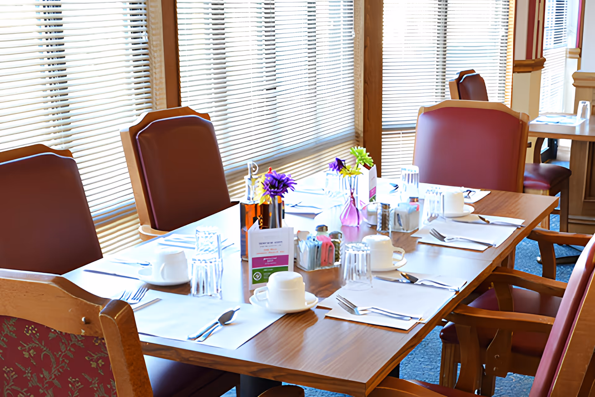 A dining table set for six with placemats, cups, glasses, cutlery, and small flower vases in a room with large windows covered by blinds. The chairs around the table have wooden frames and red cushioned seats and backs.