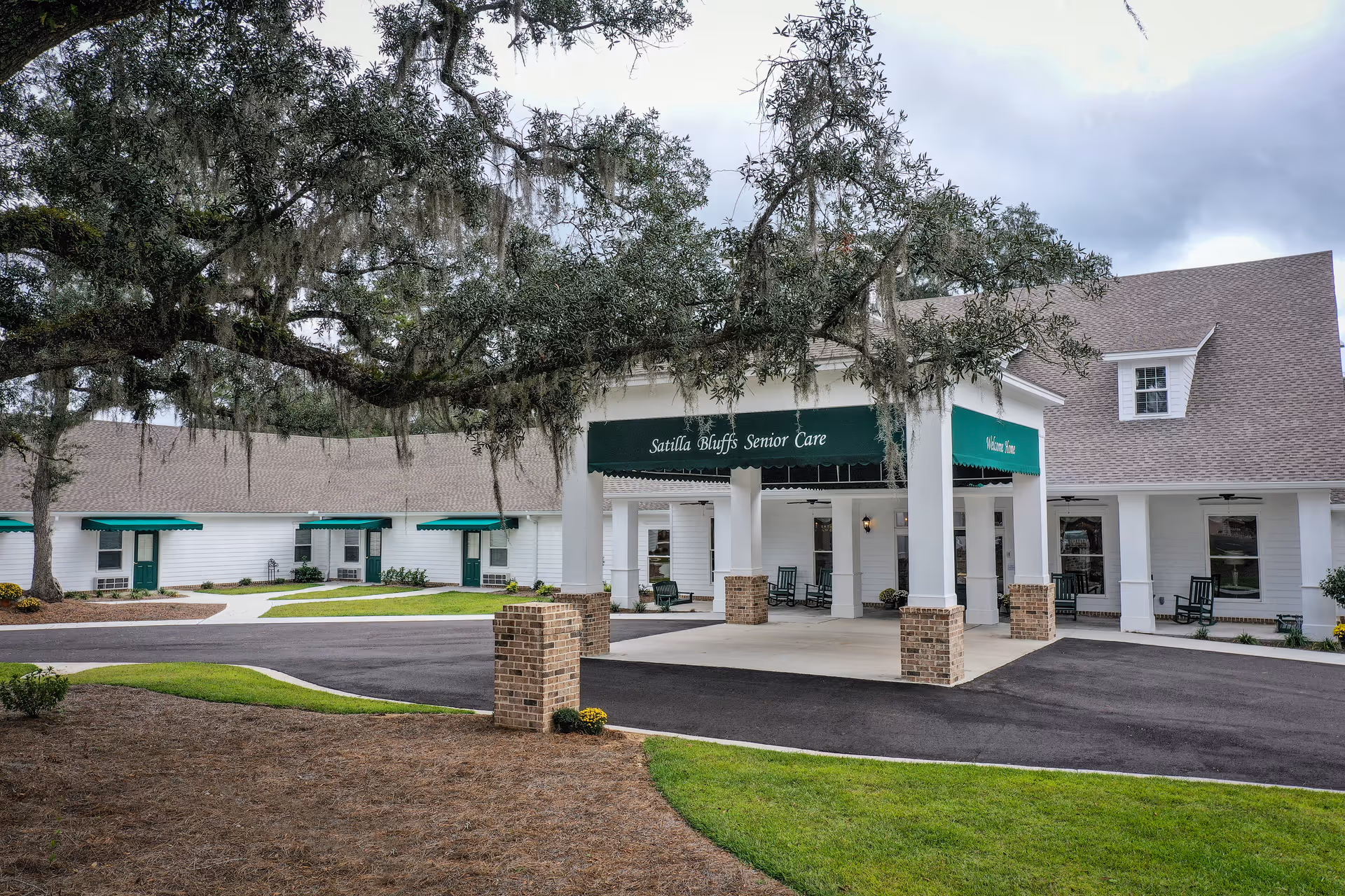 Front entrance of Satilla Bluffs Senior Care showing a covered drive-up portico with green awnings and rocking chairs in front of a white building.