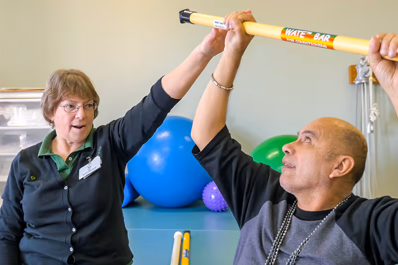 A woman and a man are engaged in a physical therapy or exercise session indoors. The woman, wearing glasses and a name tag, is assisting the man who is holding a yellow exercise bar labeled 'WATE BAR'. In the background, there are large exercise balls in blue, green, and purple colors.