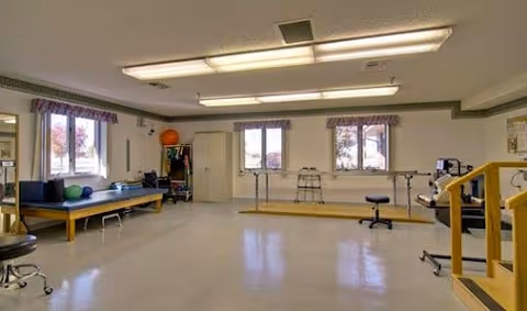 A bright therapy/exercise room with treatment tables, parallel bars, exercise balls, and seating under fluorescent lights.