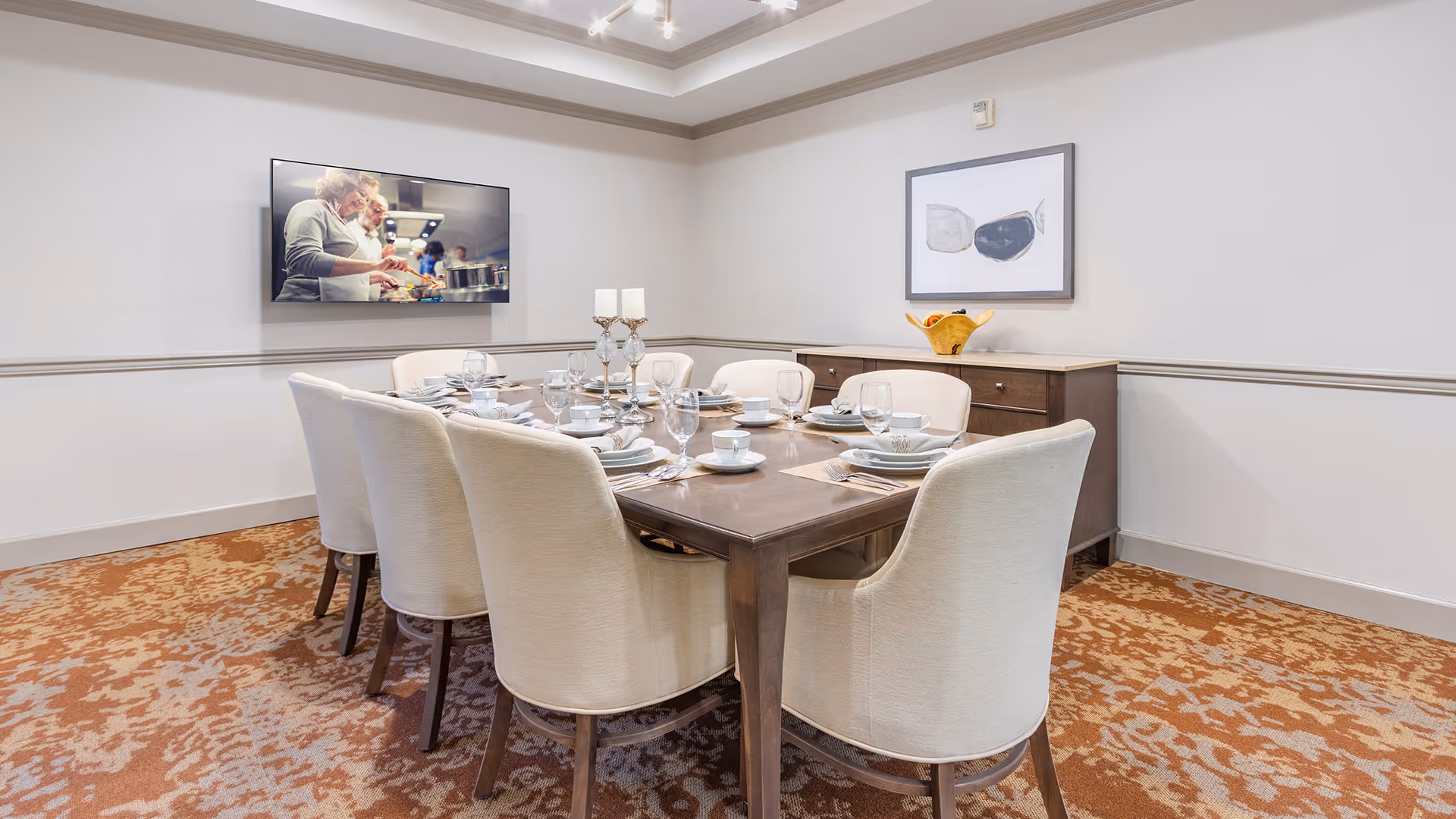 A dining room with a rectangular wooden table set for eight people with white cushioned chairs. The table is set with plates, bowls, glasses, and silverware, and has a centerpiece with three candles. On the wall, there is a flat-screen TV displaying an image of people cooking, and a framed abstract artwork above a wooden sideboard with a decorative bowl of fruit.