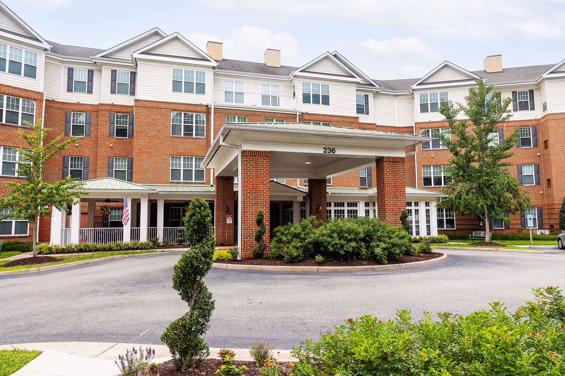 Front entrance and porte-cochere of a multi-story brick senior living building with landscaping and a circular driveway.