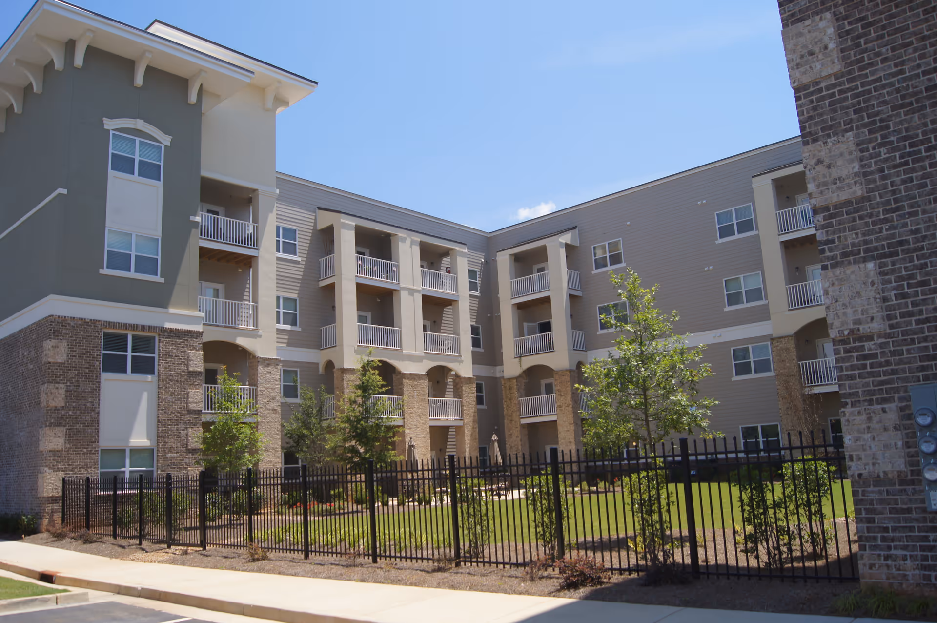Exterior view of a multi-story senior living facility building with balconies, a fenced grassy courtyard with small trees, and a clear blue sky.