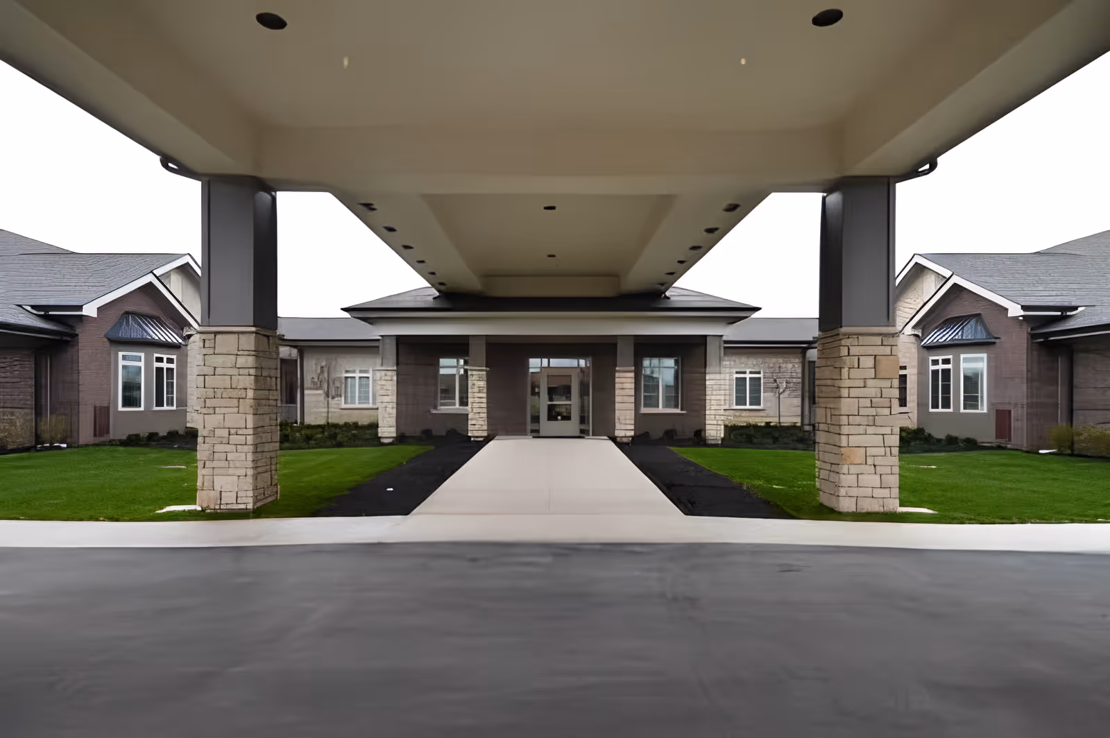 Covered entrance driveway leading to the front door of a single-story building with stone pillars and green lawns on either side.