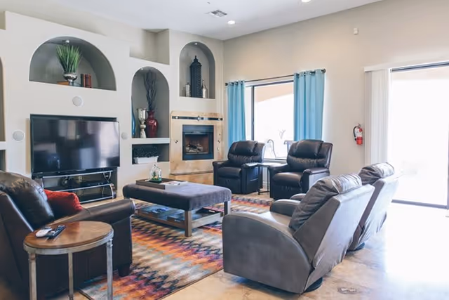 Well-lit living room with leather recliners arranged around a central ottoman, a TV, fireplace, and colorful area rug.