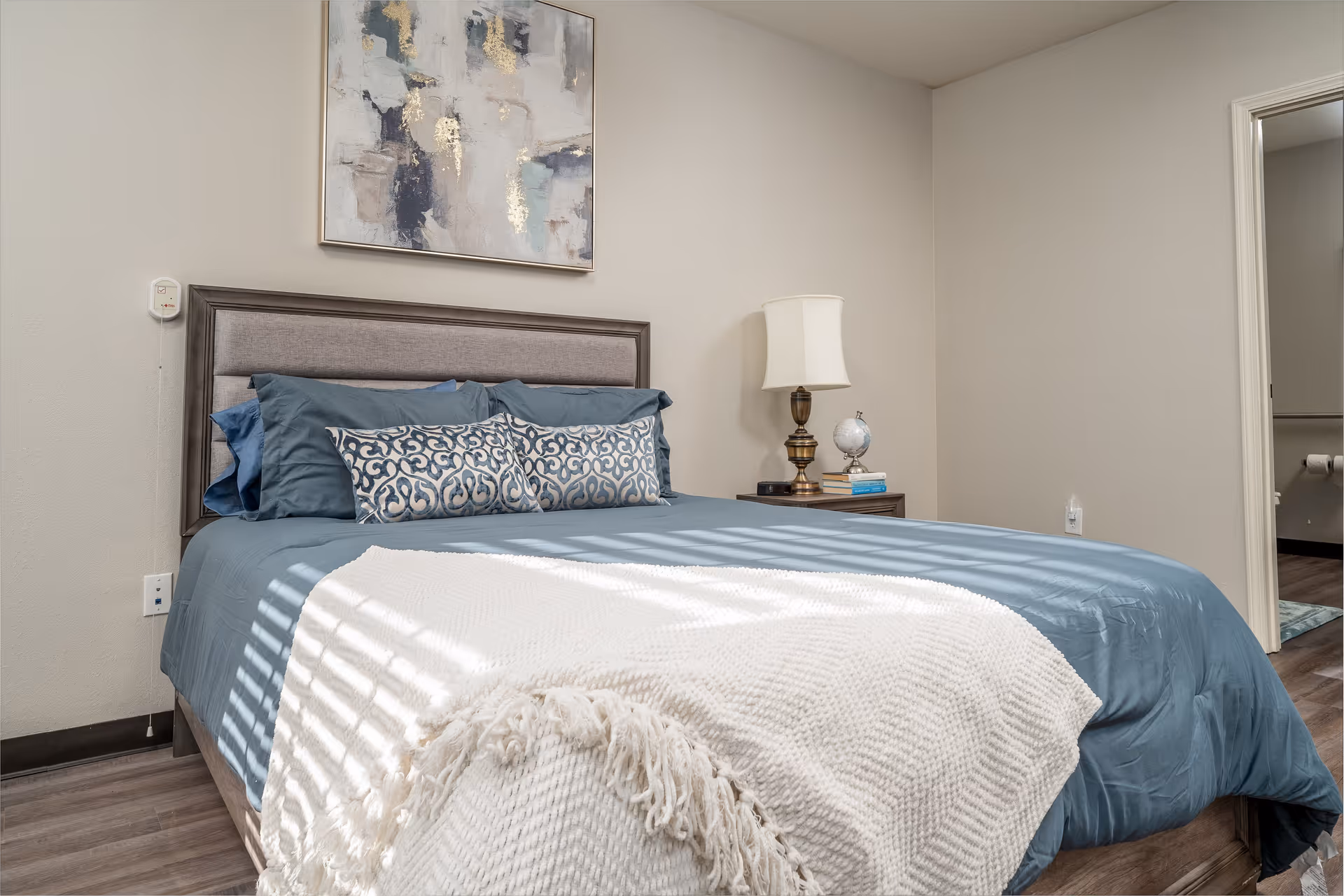 A bedroom with a neatly made bed featuring blue bedding and decorative pillows. A white textured throw blanket is draped over the foot of the bed. On the right side, there is a wooden nightstand with a brass lamp, a small globe, and a couple of books. Above the bed hangs an abstract painting with shades of blue, gray, and gold. The room has light-colored walls and wood flooring. An open doorway on the right leads to a bathroom with visible toilet paper rolls.