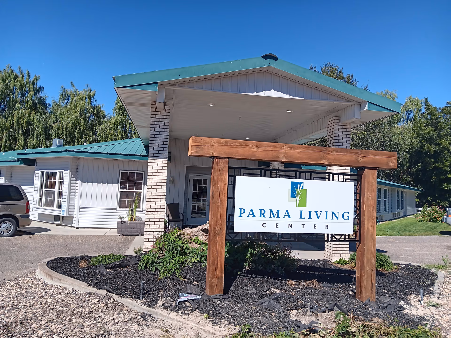 Exterior view of Parma Living Center building with a green roof and white walls. A wooden sign with the facility's name is prominently displayed in front of the entrance. There are trees and shrubs around the building under a clear blue sky.