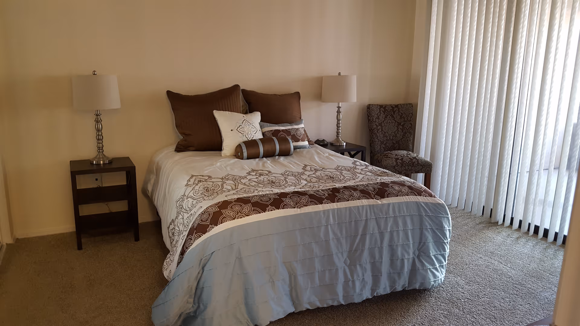 Well-lit bedroom with a made bed, two bedside tables with lamps, an upholstered chair, and vertical blinds over a sliding glass door.