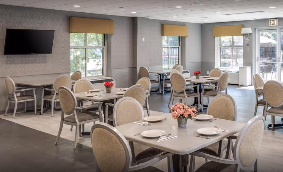 Bright communal dining room with multiple set tables and upholstered chairs, floral centerpieces, a wall-mounted TV and windows.