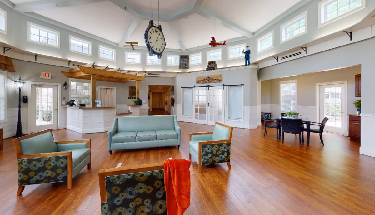 A spacious senior living facility common area with wooden flooring, light blue cushioned chairs and sofa, a reception desk with a wooden pergola above it, and large windows near the ceiling letting in natural light. The room features decorative hanging clocks and airplane models, a small table with chairs near a door, and a bright, welcoming atmosphere.