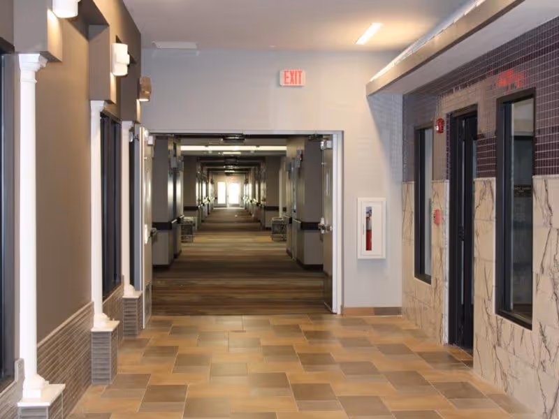 A long, well-lit hallway in a care and rehabilitation center with tiled flooring and walls partially covered with marble-like panels. The hallway has multiple doors and windows along both sides, with an exit sign visible at the far end.