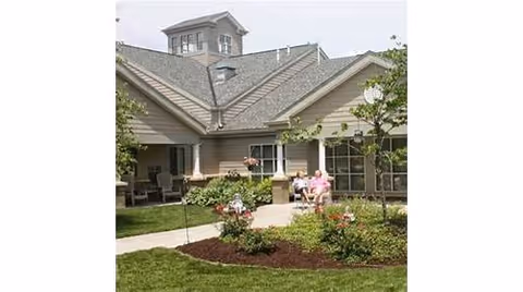 Outdoor garden area with green grass, flower beds, and a paved walkway in front of a beige building with multiple windows and a small tower-like structure on the roof. Two people are sitting on a bench near the building, enjoying the outdoor space.