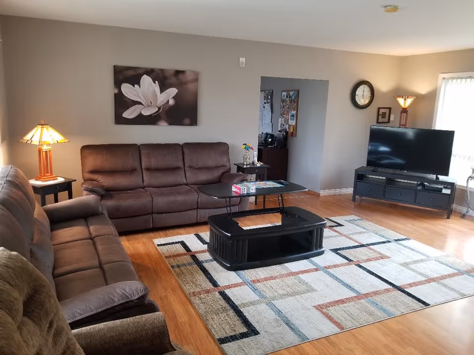 A cozy living room with two brown sofas and a brown armchair arranged around a black coffee table on a patterned area rug. There are two table lamps with warm lighting on either side of the room, a large flat-screen TV on a black TV stand, a wall clock, and a framed flower photograph on the wall. The room has wooden flooring and neutral-colored walls.
