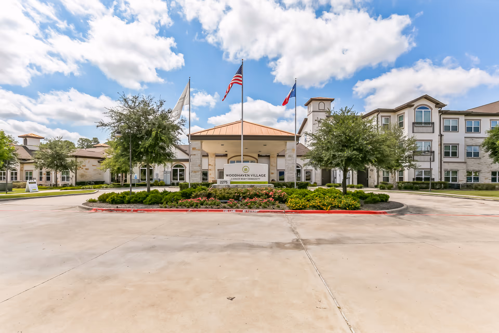 Front exterior view of Woodhaven Village senior living facility with a driveway, landscaped garden, three flagpoles with flags, and a clear blue sky with clouds.