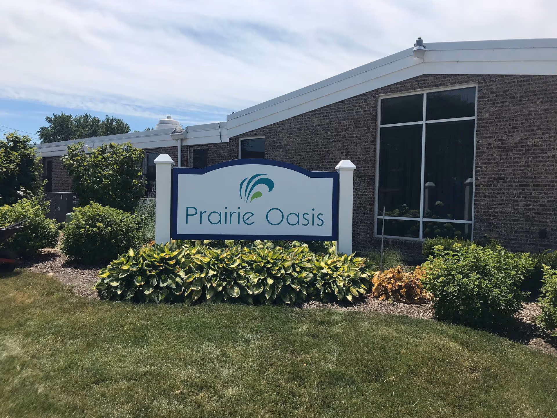 A sign reading "Prairie Oasis" sits among landscaping in front of a single-story brick building under a partly cloudy sky.