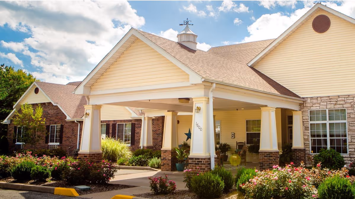 Covered portico entrance of a senior living building with white columns, brick and stone facades, and landscaped flower beds under a blue sky.