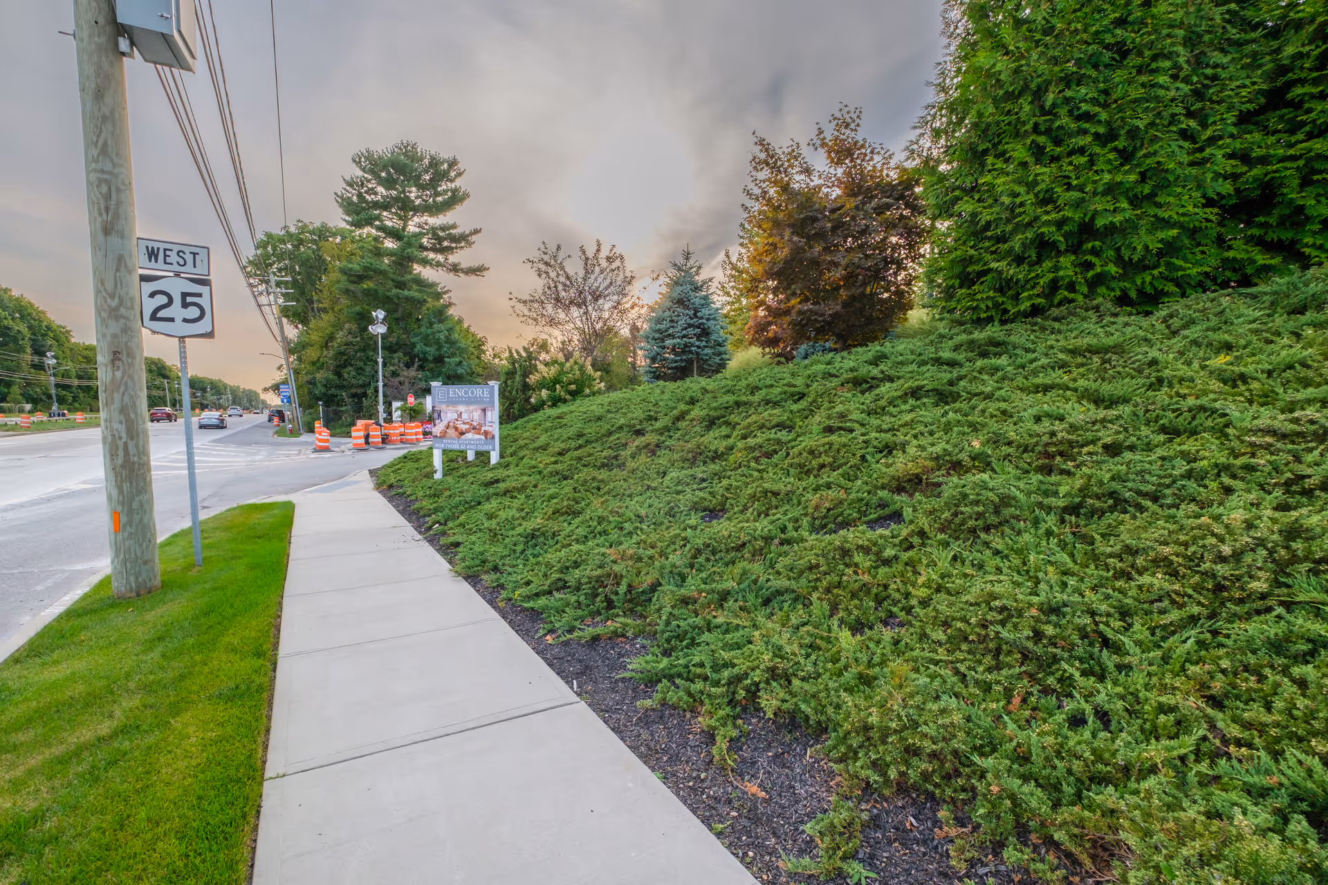 Sidewalk and roadway bordered by a landscaped grassy slope with trees and an 'Encore' sign and a West 25 road sign.
