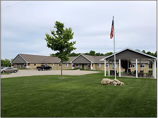 Exterior view of Maple Ridge Living Center of Lake City showing a single-story building with a covered entrance, an American flag on a flagpole, a tree, and a well-maintained lawn under a cloudy sky.
