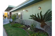 Exterior view of a single-story building with a green wall and stone accents near the base. There are outdoor wall lights mounted on the building, a landscaped garden with various plants and shrubs, and a well-maintained lawn. A white fence is visible on the left side of the image.