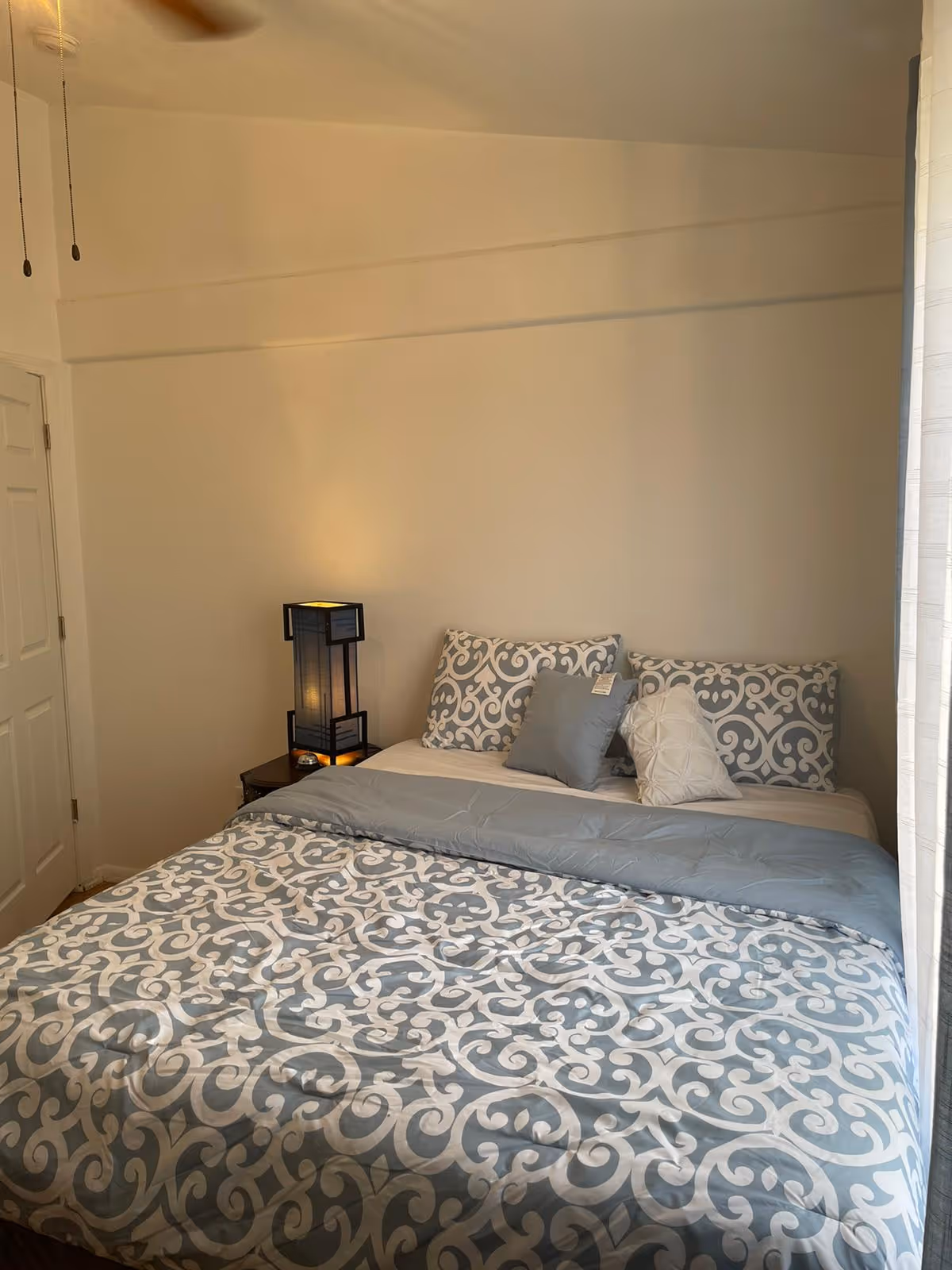 A bedroom with a neatly made bed featuring a blue and white patterned comforter and matching pillows. There is a small bedside table with a modern lamp emitting a warm light. The walls are plain and light-colored, and a ceiling fan is partially visible at the top left corner.