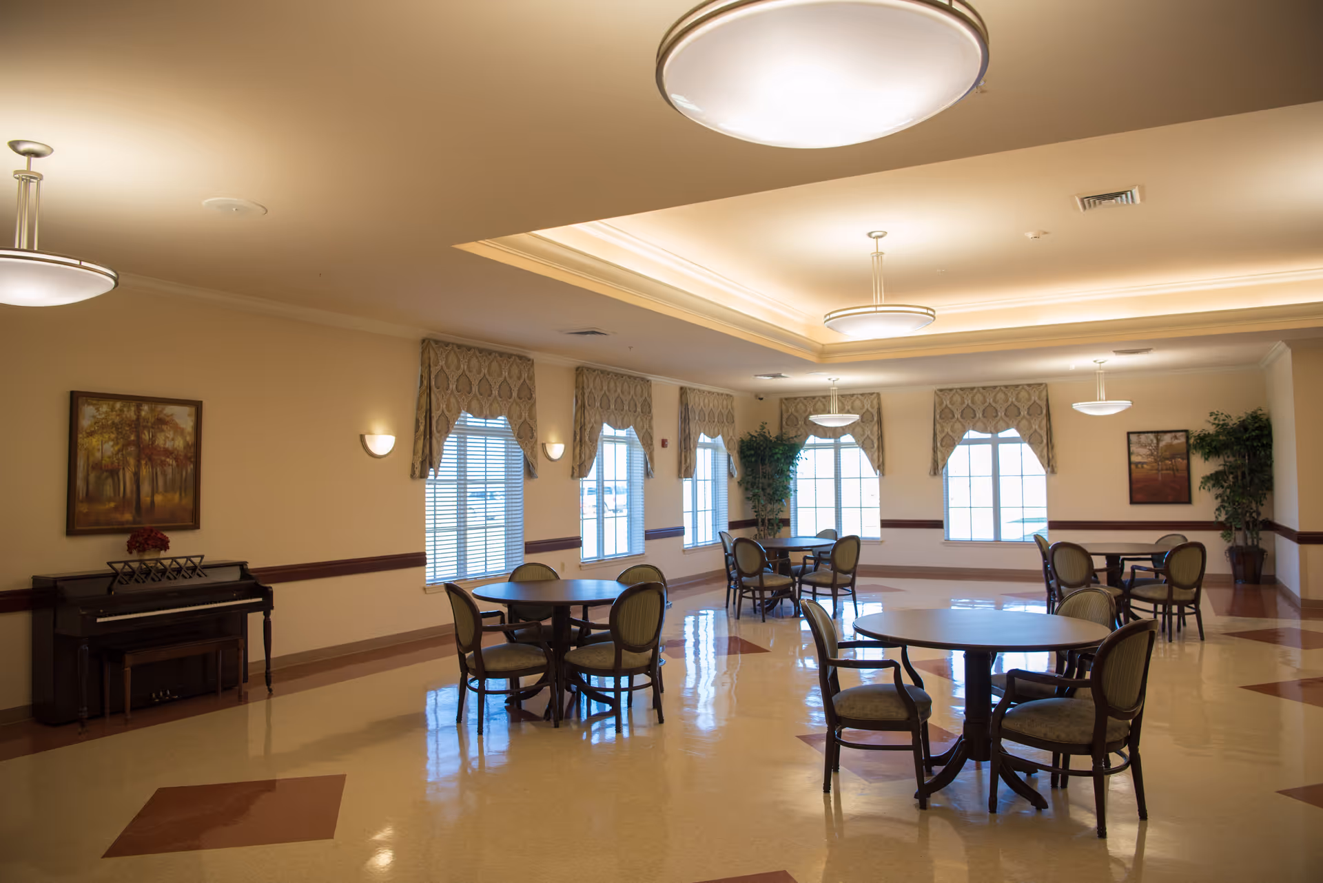 A spacious, well-lit room with round tables and chairs arranged for dining or socializing. The room features large windows with patterned valances, ceiling lights, framed artwork on the walls, a piano with a flower arrangement on top, and potted plants in the corners.