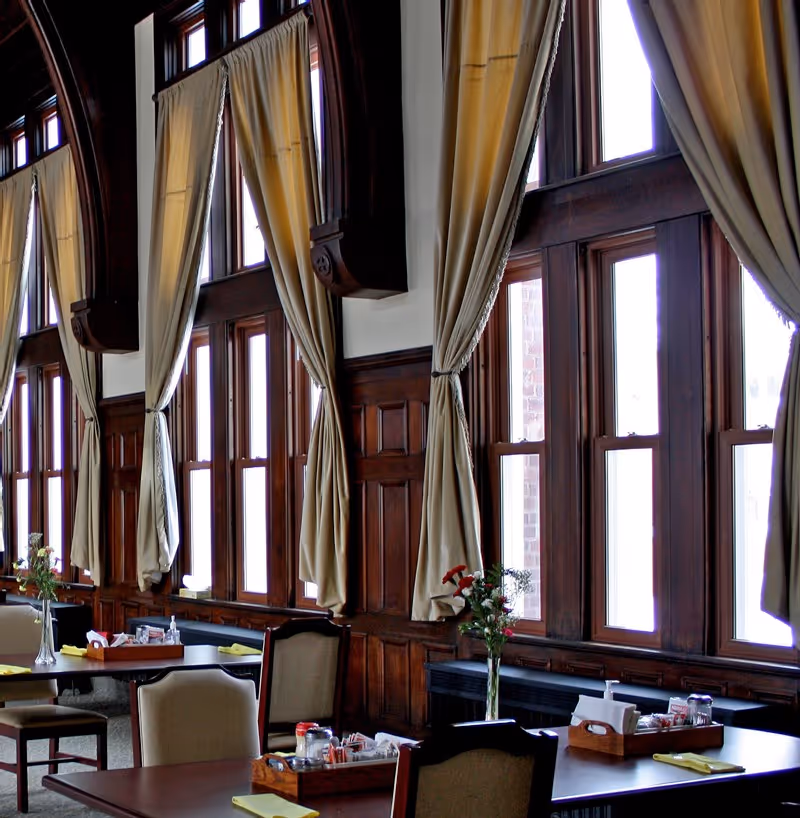 Dining area with multiple tables and chairs, each table set with napkins, condiments, and a vase with flowers. Large wooden framed windows with beige curtains tied back, allowing natural light to fill the room. The walls have dark wood paneling.