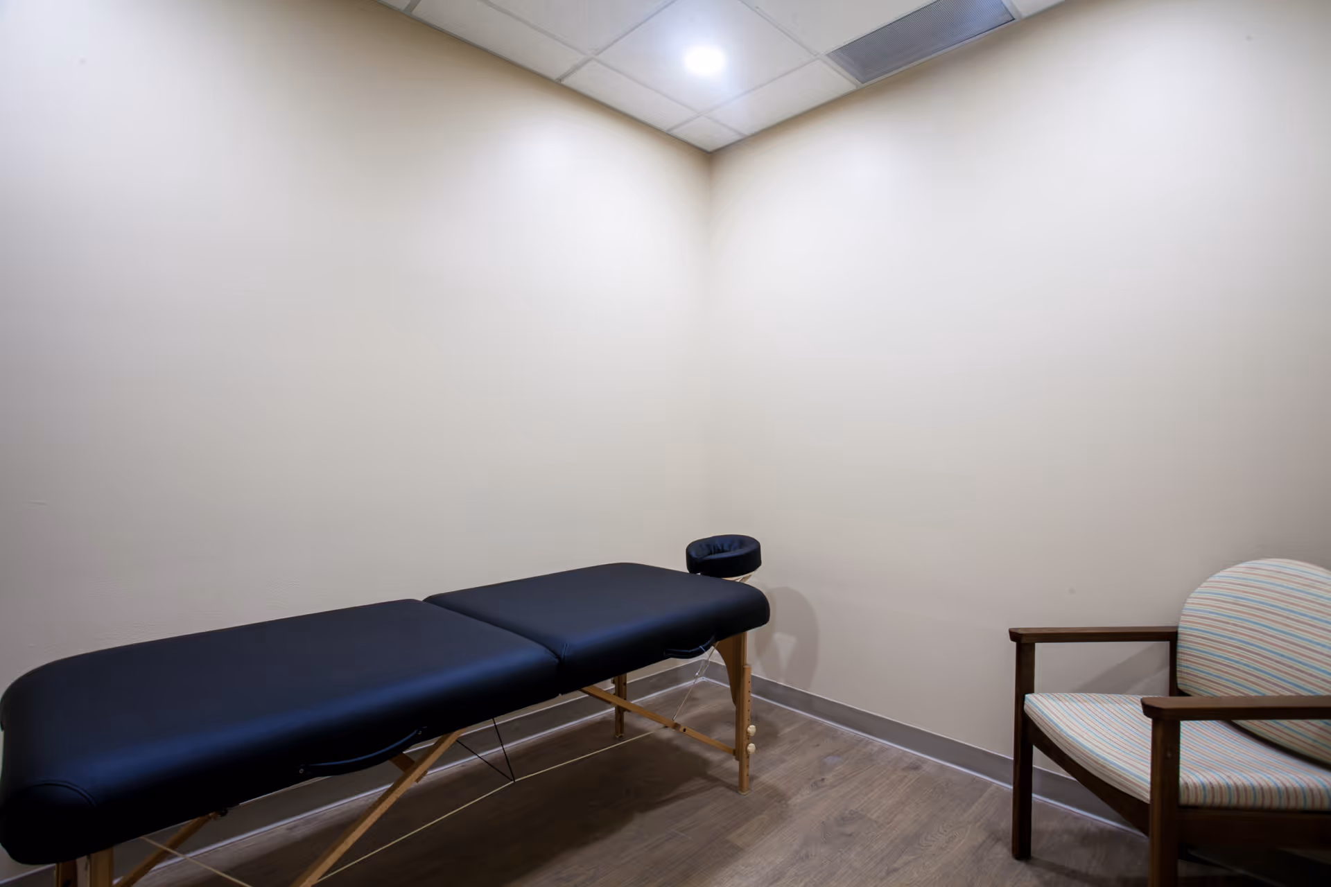 A small room with beige walls and wood flooring featuring a black massage table and a wooden chair with striped upholstery. The ceiling has recessed lighting.