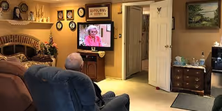 An elderly man sitting in a recliner chair watching television in a cozy living room with beige walls, a fireplace with decorative items on the mantel, several framed pictures and clocks on the walls, a wooden cabinet with a TV, and a small dresser with various items on top.