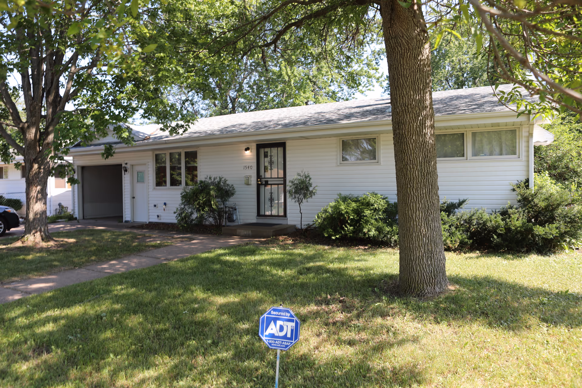 Single-story white house with a front door, several windows, a garage, and a large tree in the front yard. The lawn is green with some bushes near the house and an ADT security sign is placed on the grass near the sidewalk.