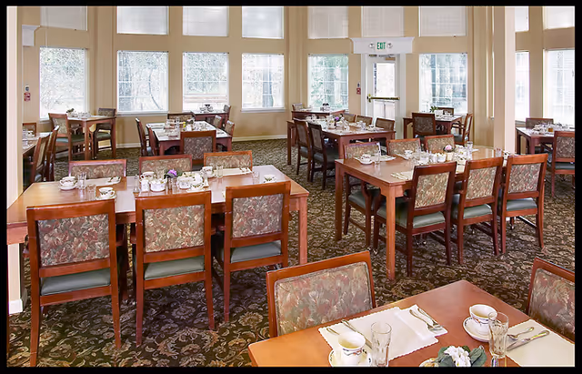 Bright dining room with multiple wooden tables and chairs set for meals and large windows along the back wall.