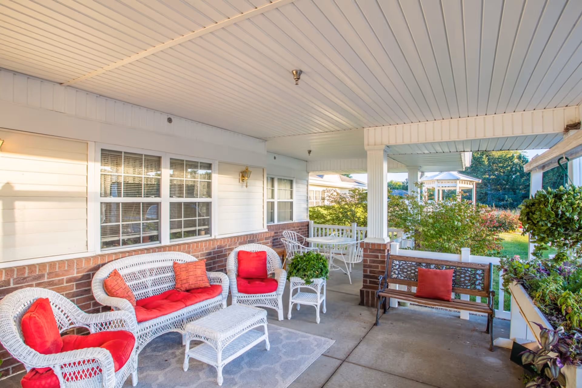 Covered porch with white wicker seating and red cushions, a bench, table, and potted plants overlooking a garden.