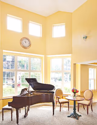 Bright room with large windows letting in natural light, featuring a grand piano, a small round table with a vase of flowers, and two upholstered chairs. A clock is mounted on the wall above the windows.