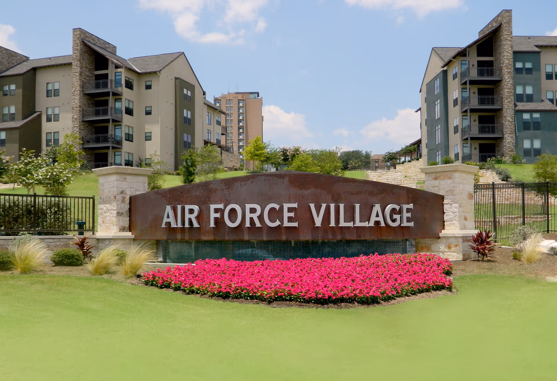Entrance sign for Air Force Village with modern apartment buildings in the background, green grass, and bright pink flowers in front of the sign under a partly cloudy sky.