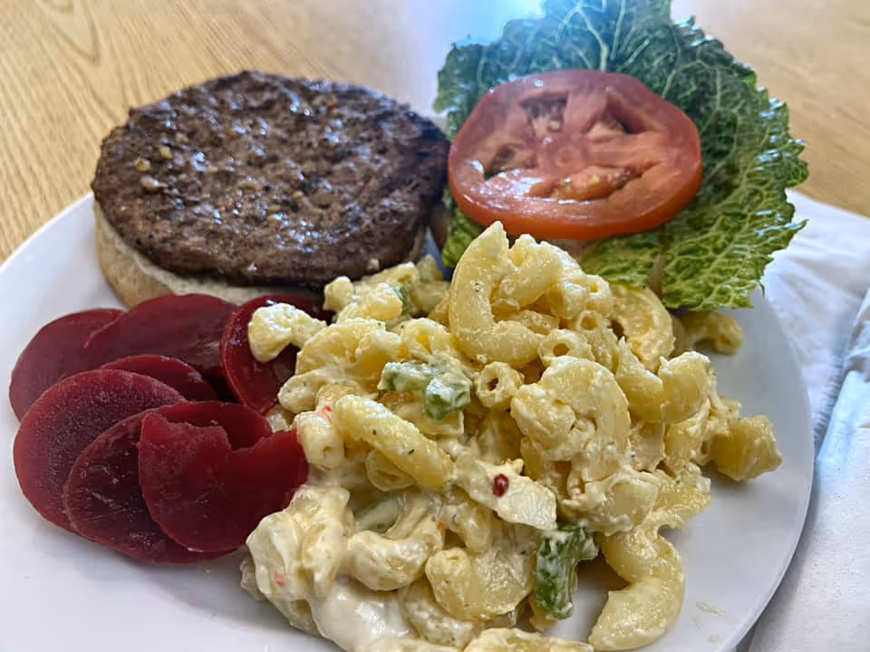 A plate of food featuring a hamburger patty on a bun with lettuce and tomato, macaroni salad with vegetables, and sliced beets, placed on a wooden table.