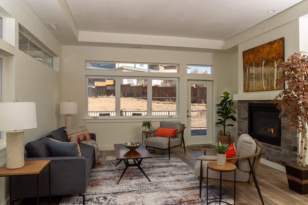 A cozy living room with a dark gray sofa, two light gray armchairs with red cushions, a wooden coffee table, and side tables with lamps and plants. There is a fireplace with a stone surround and a painting of autumn trees above it. Large windows and a glass door let in natural light and show a fenced backyard.