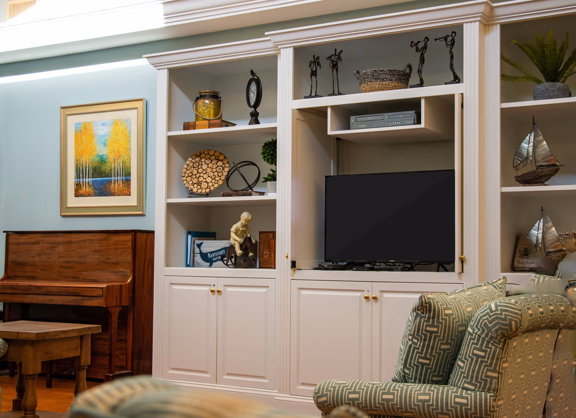 A cozy living room area with a wooden piano and a small wooden table on the left. A white built-in shelving unit holds decorative items including sculptures, a clock, a basket, and a flat-screen TV. A patterned armchair is partially visible in the foreground. A framed painting of yellow trees by a lake hangs on the light blue wall above the piano.