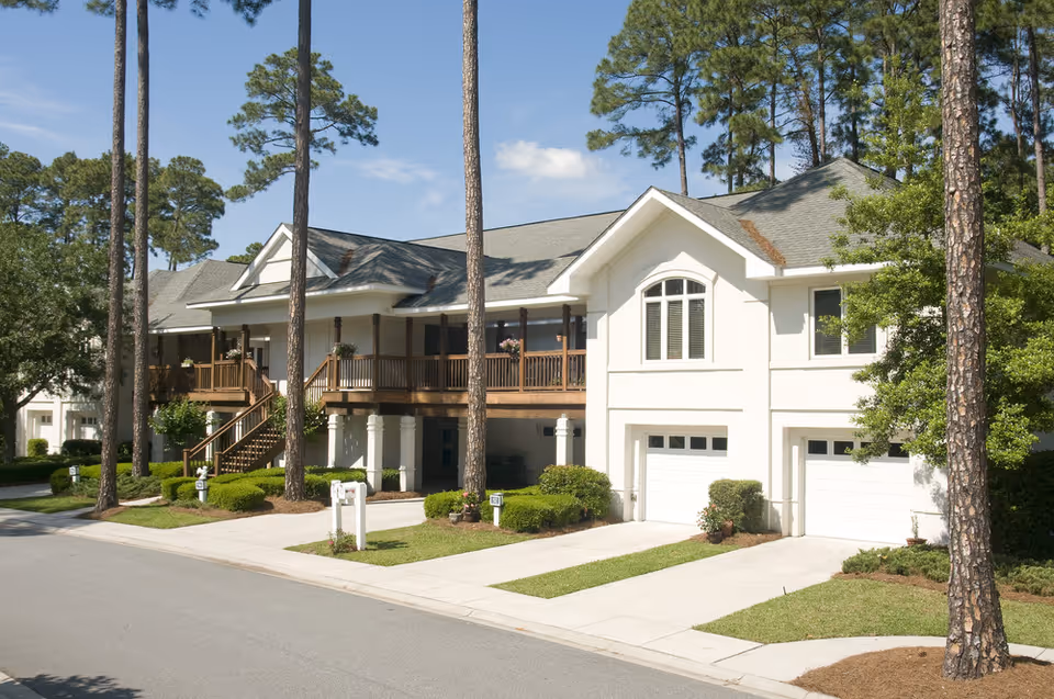 White multi-unit senior living building with garages, balconies, and tall pine trees along a street.