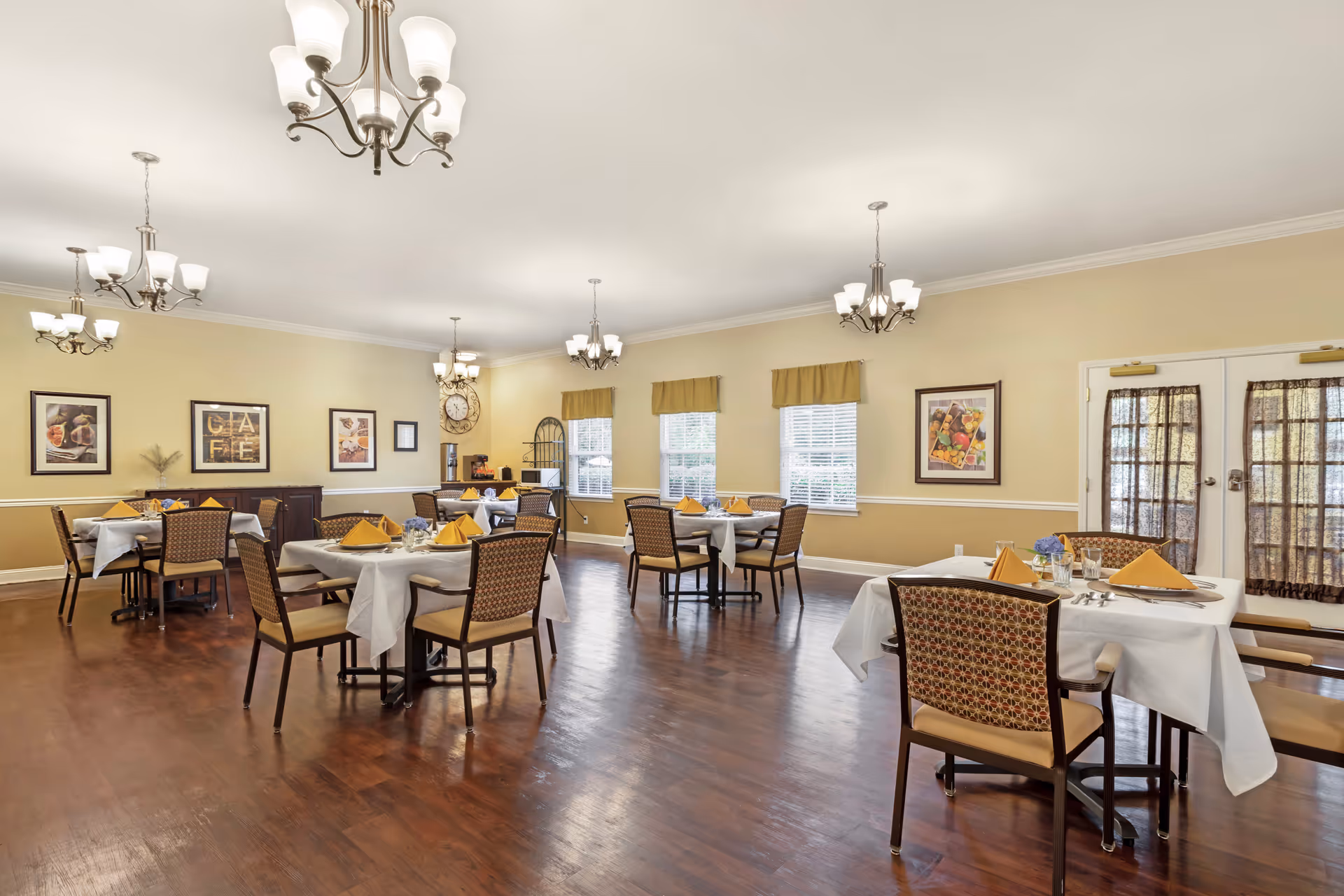A spacious dining room with several tables covered in white tablecloths, each set with yellow folded napkins, glasses, and silverware. The room has wooden flooring, beige walls with framed artwork, multiple windows with valances, and several ceiling light fixtures. There is a coffee station and a large decorative clock on one wall.