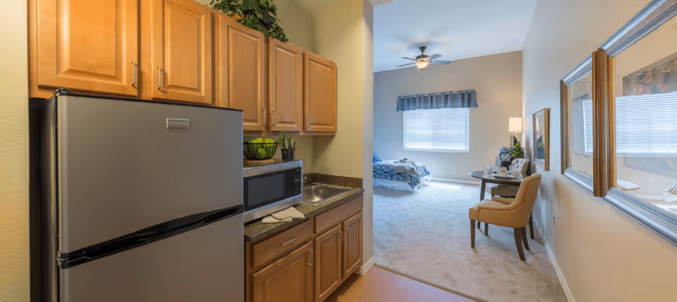 Small kitchenette with a refrigerator, microwave and sink opening into a carpeted studio living/bedroom area with a window and chair.