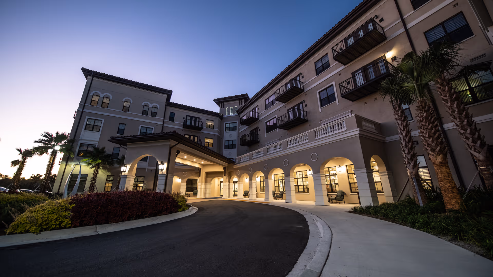 Exterior view of a multi-story senior living facility building at dusk with arched entryway, balconies, palm trees, and landscaped bushes along a curved driveway.