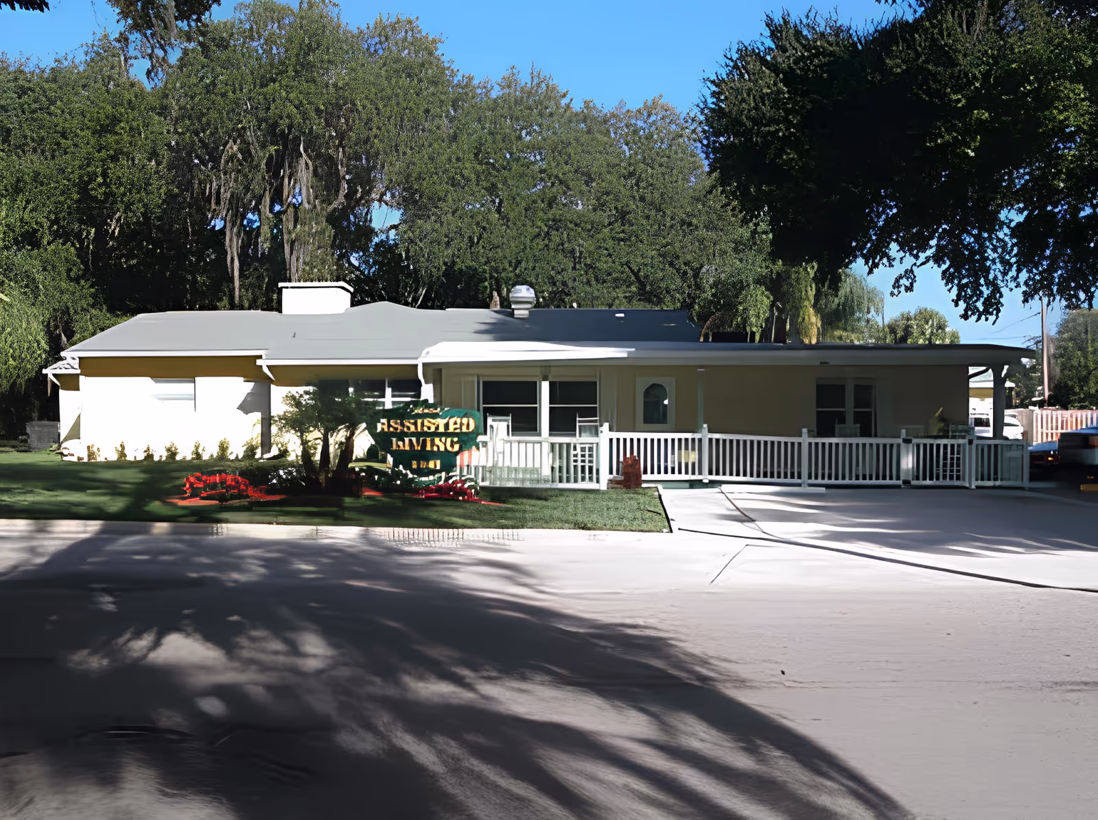 Single-story building with a light-colored exterior surrounded by green trees and a well-maintained lawn. A sign in front reads 'Assisted Living'. The building has a white railing along a small porch area and a driveway in front.