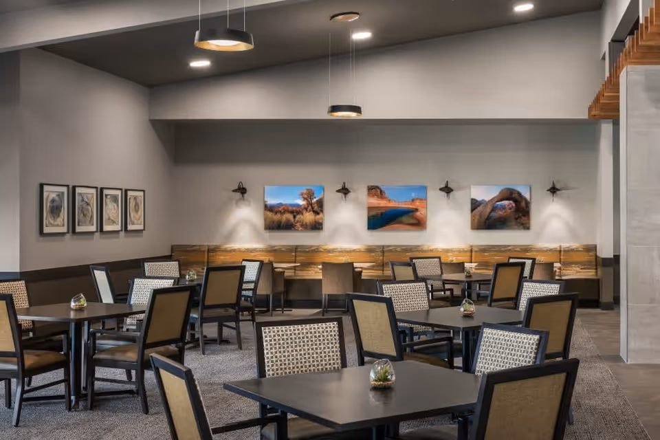 A modern dining room with multiple square tables and cushioned chairs arranged neatly. The walls are decorated with framed artwork and landscape photographs. Pendant lights hang from the ceiling, illuminating the space with a warm ambiance.