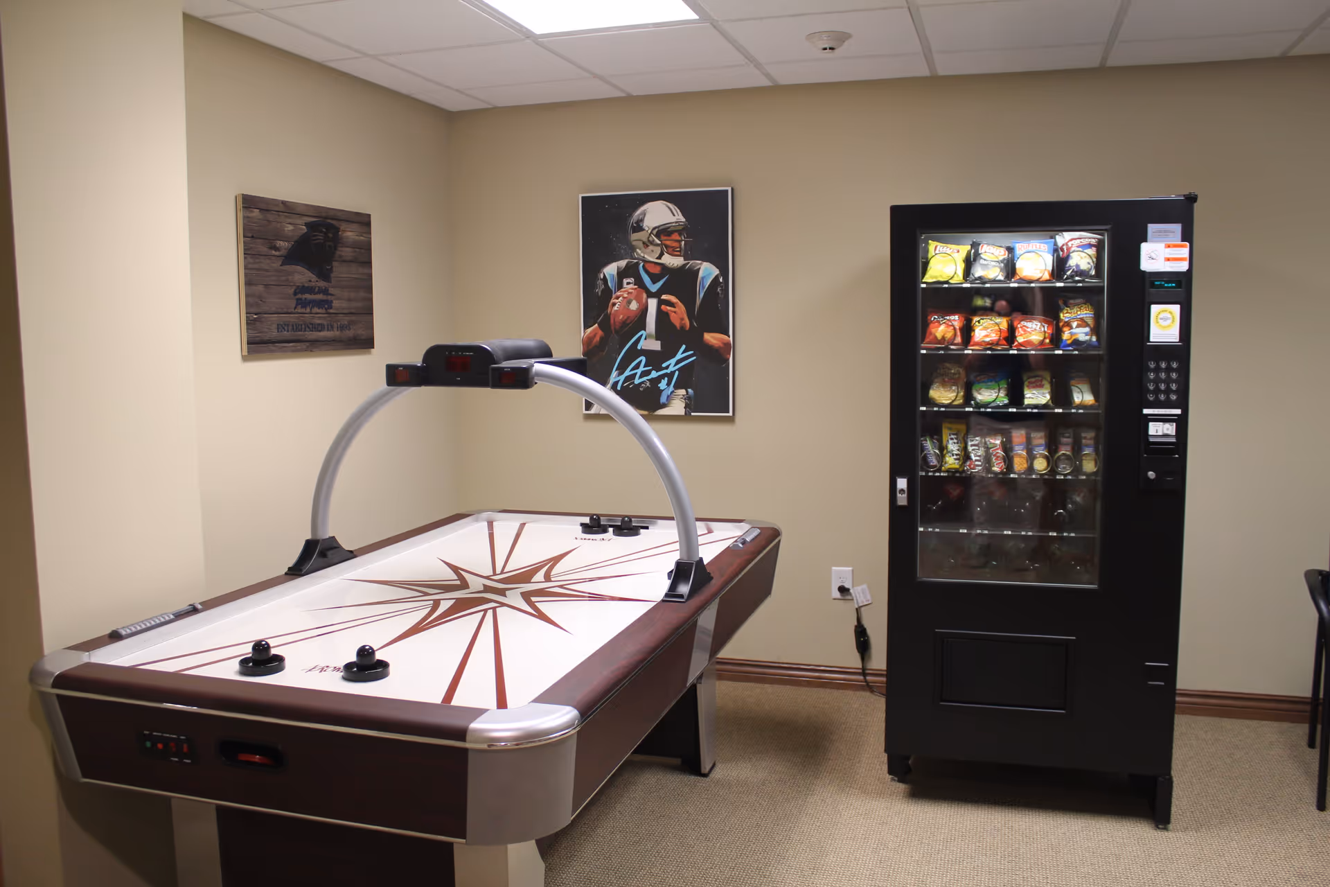 A small recreational room with an air hockey table and a vending machine stocked with snacks. The walls are beige, and there are two framed pictures on the wall, one featuring a football player and the other a sports team logo.