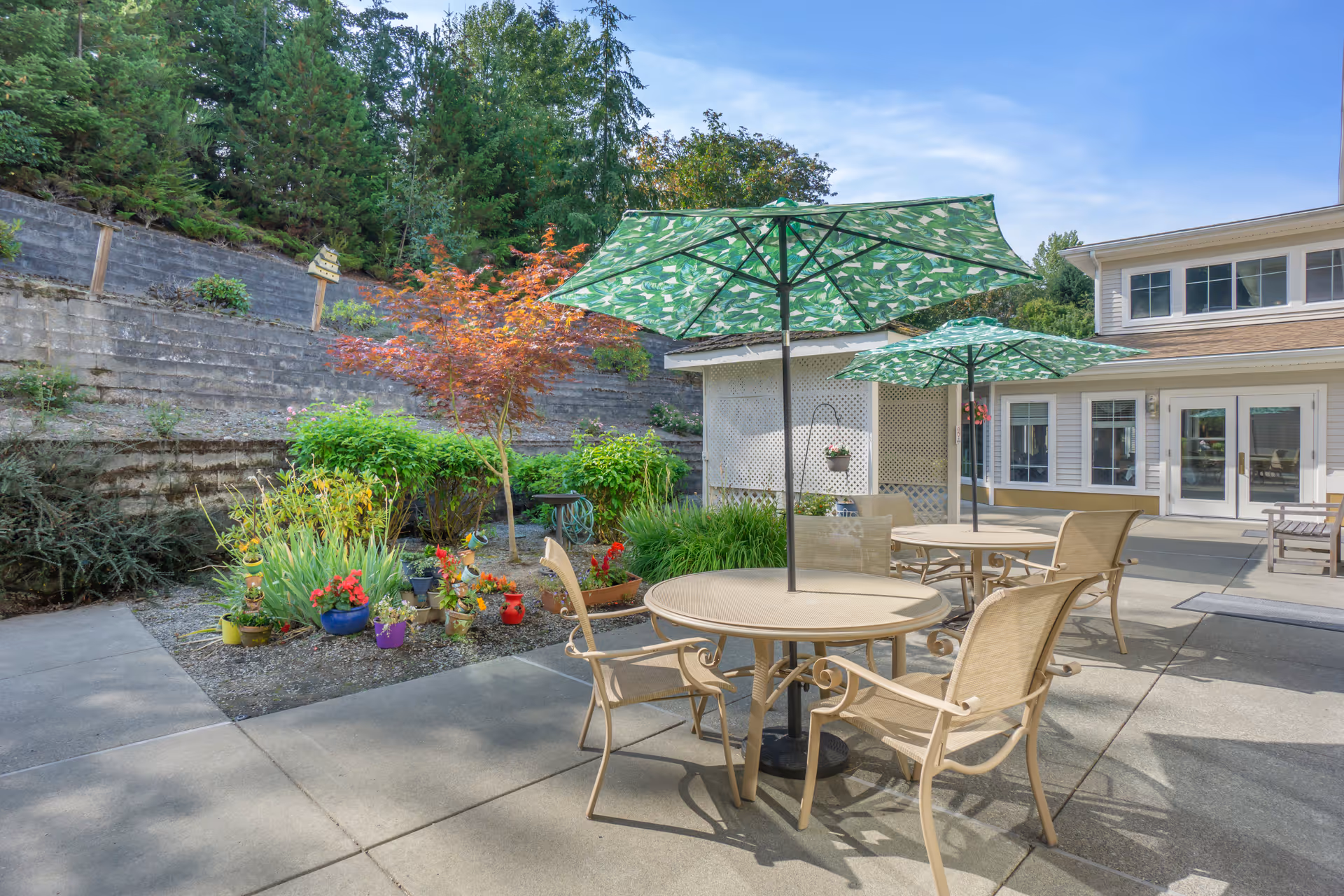 Outdoor patio with round tables, chairs and green umbrellas beside a garden and a building entrance.