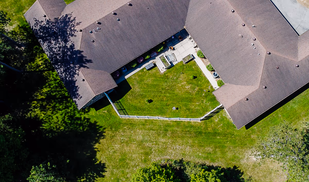 Aerial view of a senior living facility building with a fenced green courtyard in the center. The building has a brown roof and is surrounded by grass and trees casting shadows.