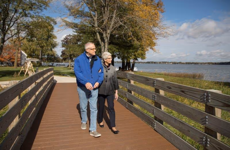 An elderly couple walking arm in arm on a wooden boardwalk beside a lake, surrounded by trees with autumn foliage under a partly cloudy sky.