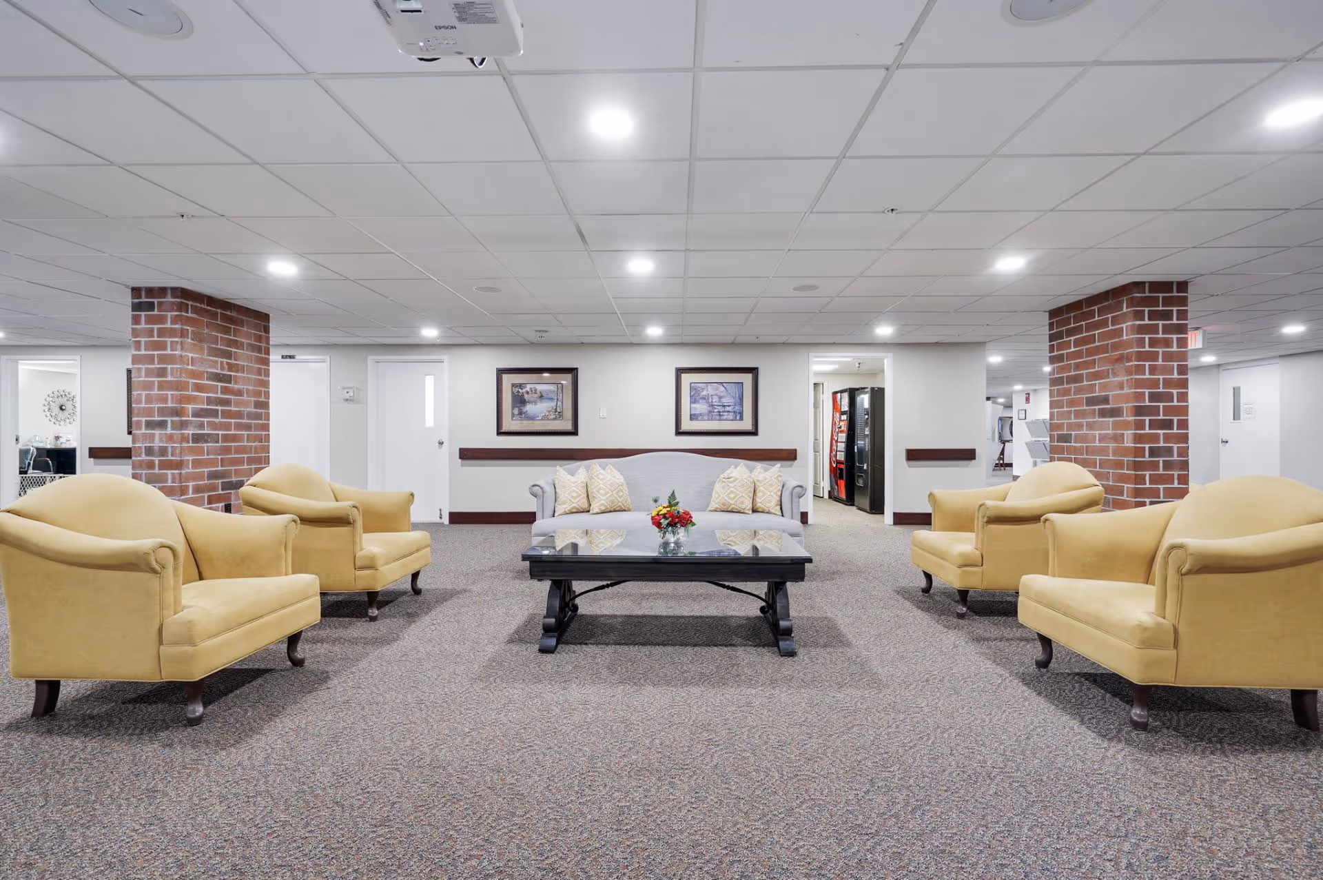 A spacious lounge area with a gray sofa and four yellow armchairs arranged around a black coffee table with a small flower arrangement. The room has carpeted floors, white walls, two framed pictures on the wall, and exposed brick columns. Ceiling lights illuminate the space, and vending machines are visible in the background.