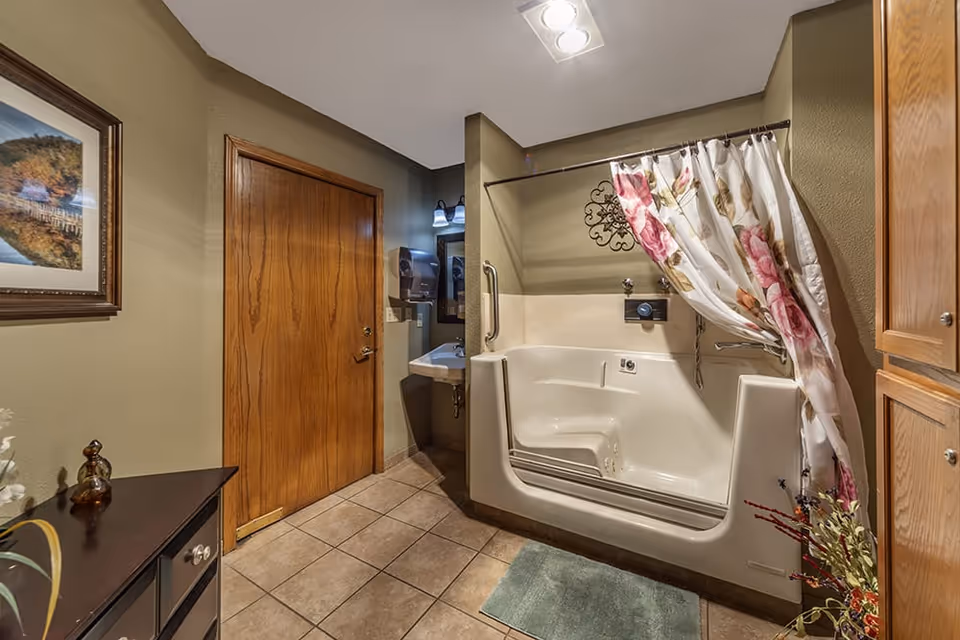 A bathroom with a walk-in bathtub featuring a floral shower curtain, a small sink with a mirror and light above it, a wooden door, tiled floor, and a cabinet on the right side. There is a framed picture on the left wall and a small rug in front of the bathtub.